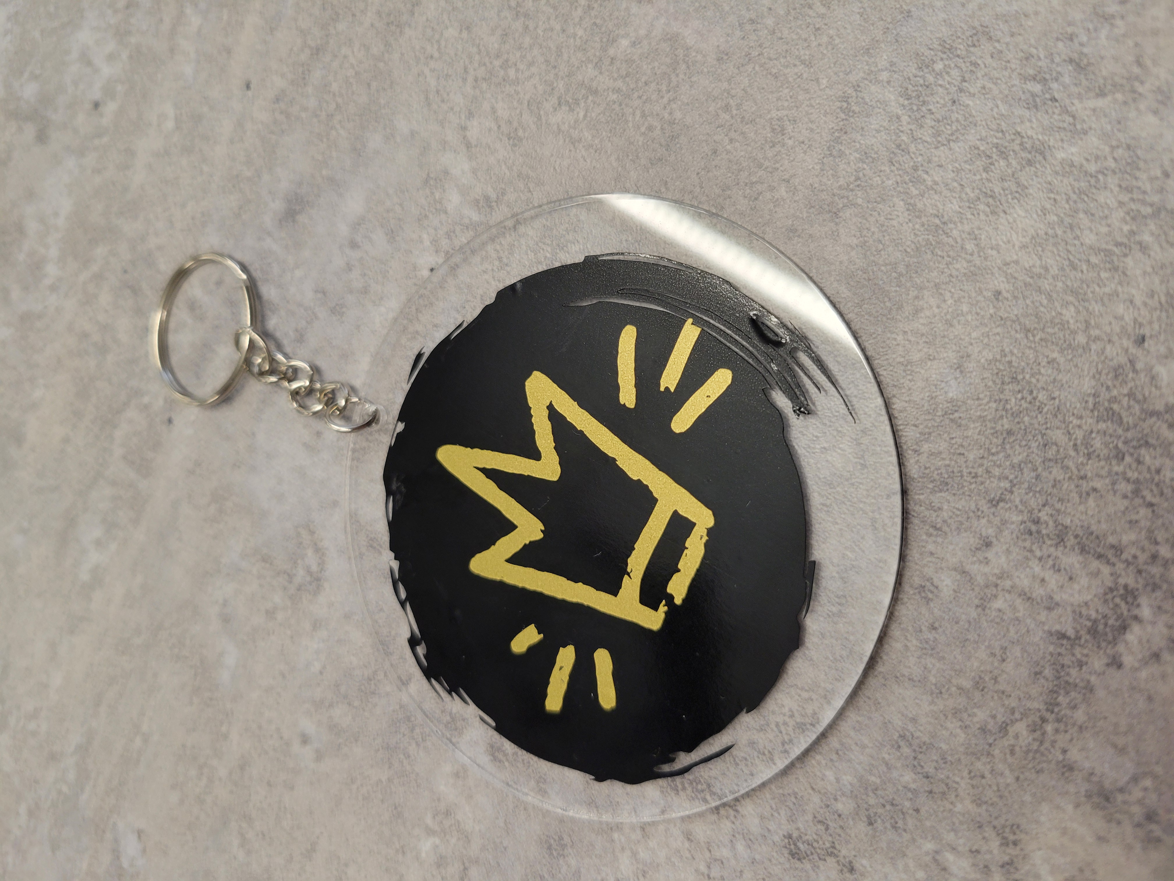 an acrylic keychain with a gold crown against a black brush backdrop